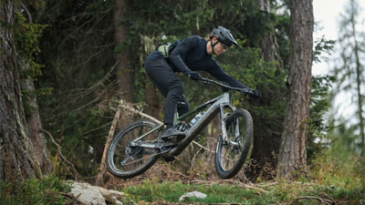 Zu sehen ist ein Mann auf einem grauen Cube Stereo Hybrid E-Bike, welcher über einen Hang im Wald springt.