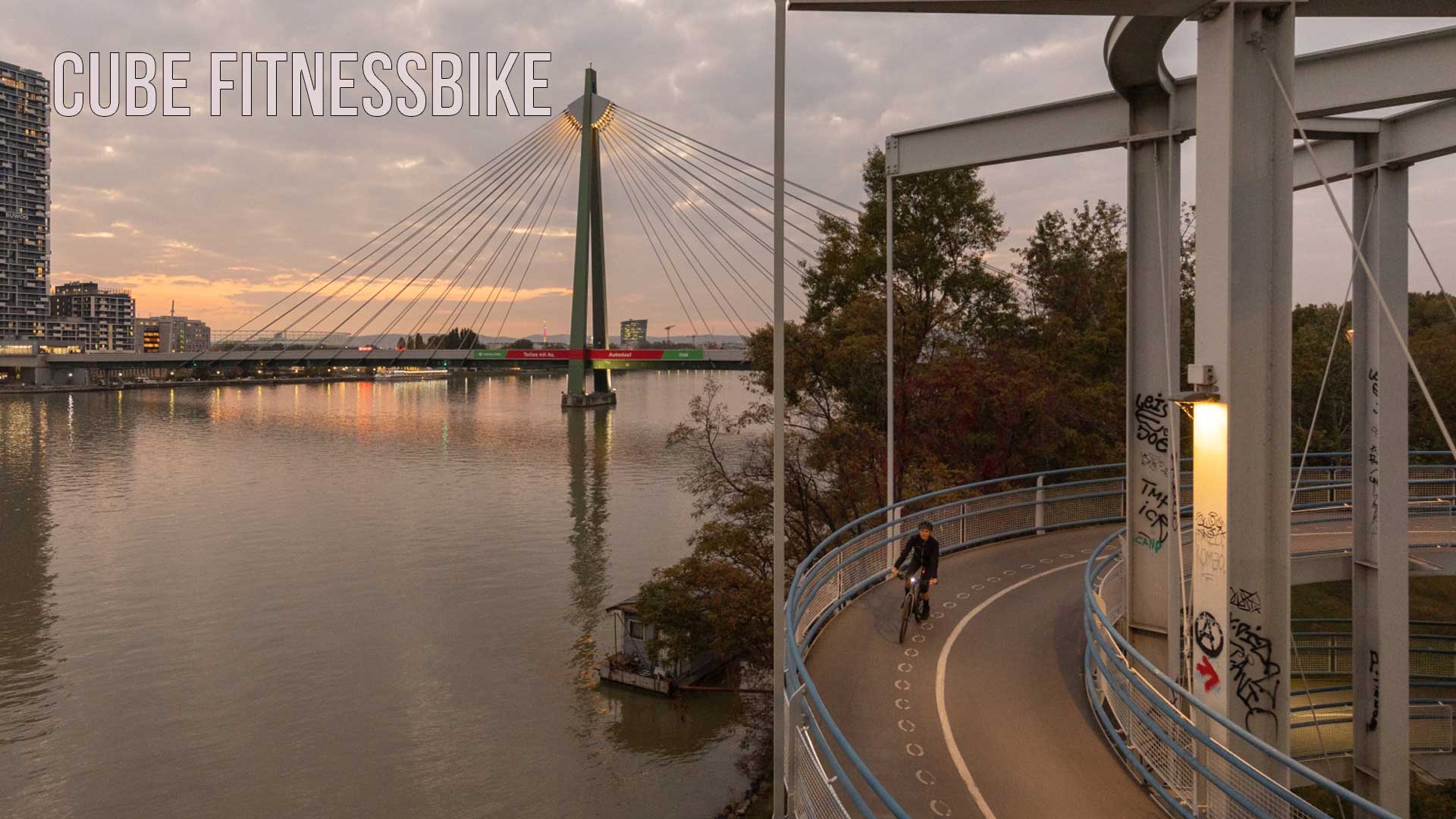 Zu sehen ist eine Stadt am Fluss, Im Vordergrund ist eine Radfahrerin, welche bei Dämmerung auf dem Radweg unterwegs ist. Das Bild ist mit dem Titel Cube Fitnessbike beschriftet.