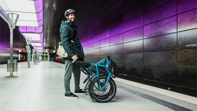 Zu sehen ist ein Mann, der an einer U-Bahn Station steht. Vor ihm steht ein eingeklapptes blaues Faltrad.
