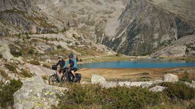 Ein Paar fährt mit dem Trekkingräd durch steinige Landschaft, im Hintergrund sind Berge und ein See zusehen.