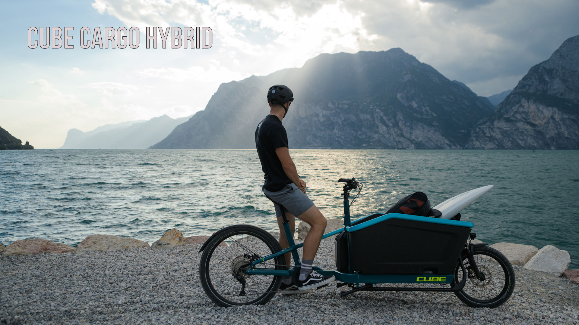 Zu sehen ist ein Mann auf einem blauen Lastenrad, er steht am Ufer eines Sees und betrachtet die Berge im Hintergrung. Beschrieben ist das Bild mit dem Titel Cube Cargo Hybrid.