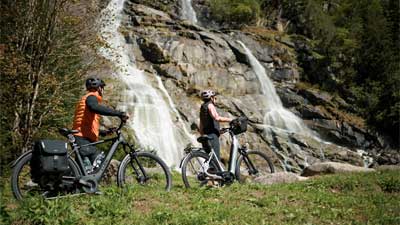 Ein Paar schiebt seine Fahrräder über eine Wiese, im Hintergrund ist ein Wasserfall.